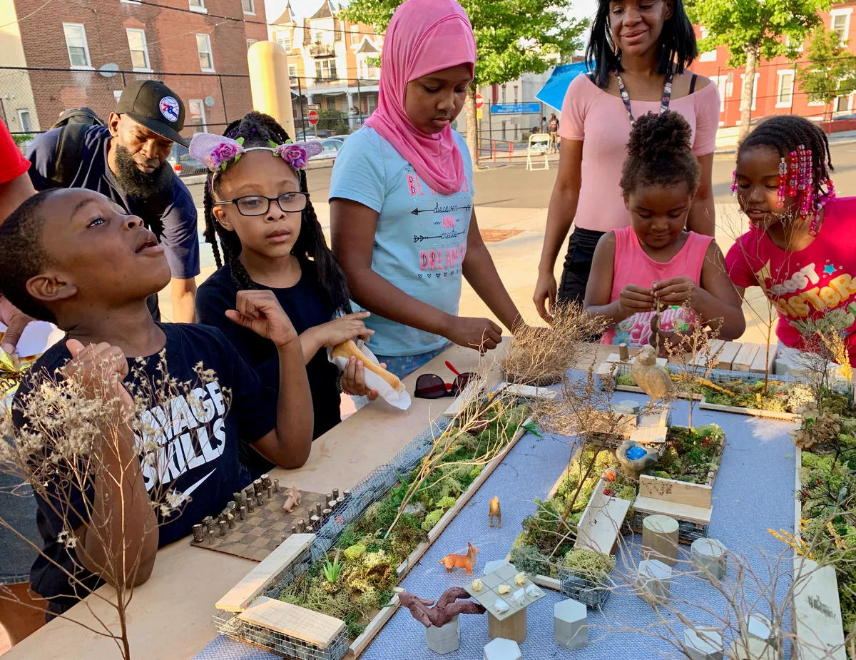 A group of children and adults gathered around a park-piece model on build day at Melon Street Park