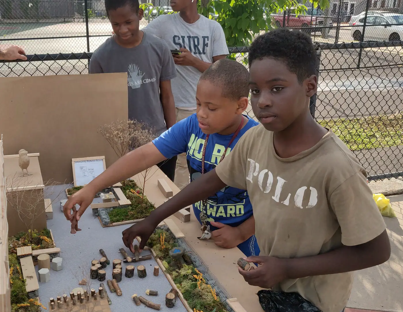 Teenagers gather around an outdoor model-making table, arranging small sticks and a model birdhouse on a tabletop park model behind a chain-link fence.