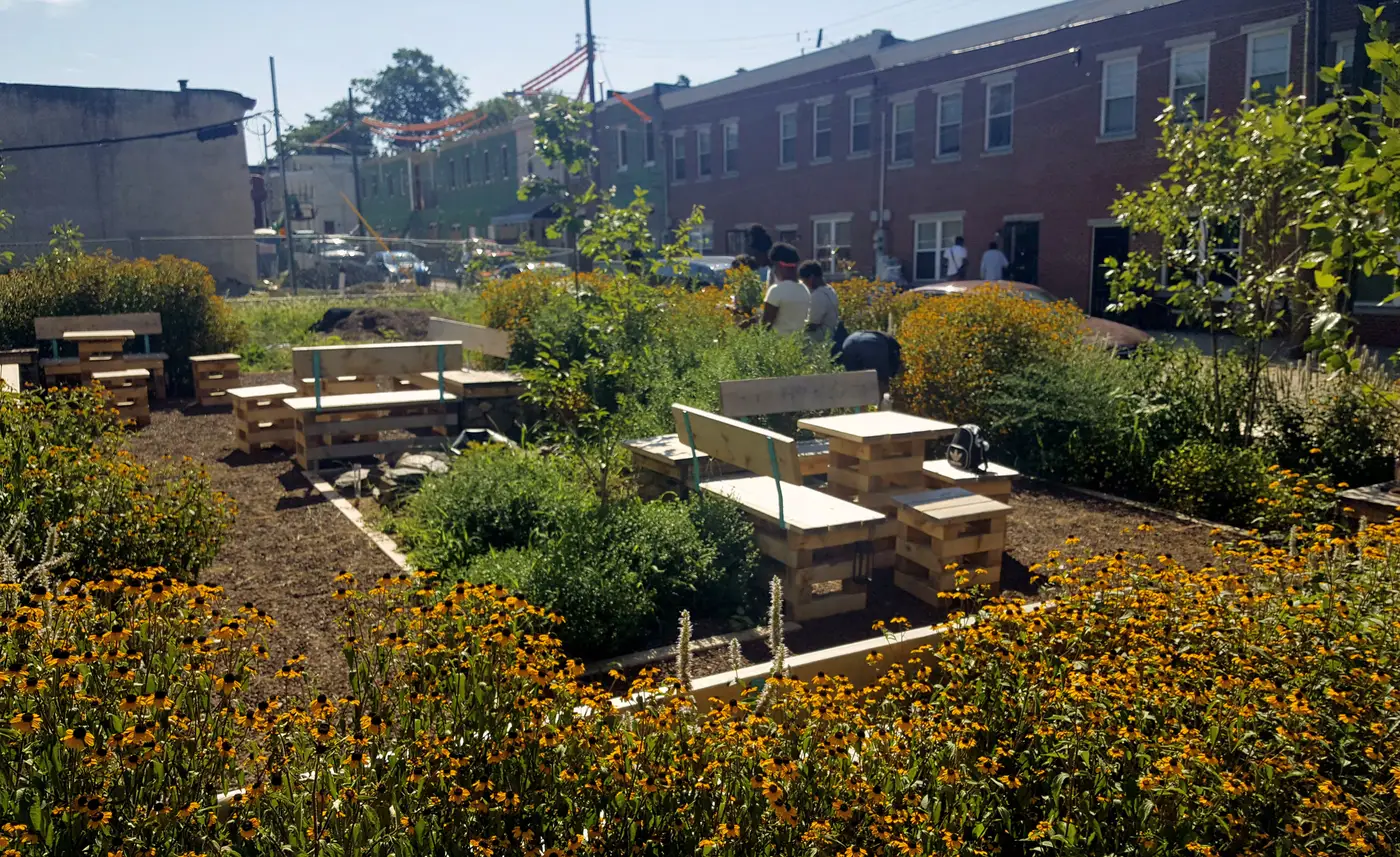 Neighbors gathered among wooden benches and tables at the Melon Street Park, a Park in a Truck park built on a formerly vacant lot in Mantua, Philadelphia, surrounded by black-eyed Susans and rowhomes.